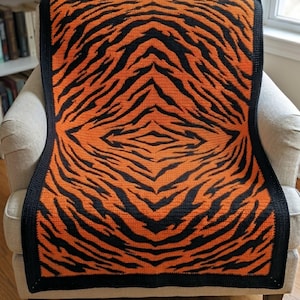 Puede incluir: Una manta de ganchillo con un diseño de rayas de tigre en naranja y negro. La manta está sobre un sillón beige. Los bordes de la manta tienen un borde negro. El diseño es simétrico, con las rayas convergiendo hacia el centro.