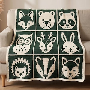 May include: A rectangular crochet blanket featuring a grid of nine square panels, each displaying a different forest animal face in cream against a dark forest green background. The animals include a fox, bear, raccoon, owl, deer, rabbit, hedgehog, badger, and cat.