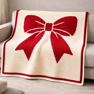 Könnte beinhalten: Eine cremefarbene gehäkelte Decke mit einem großen roten Schleifenmuster. Die Decke hat einen roten Rand und ist über einem grauen Sofa drapiert. Die Schleife ist ein herausragendes Merkmal und verleiht der gemütlichen Wohnkultur eine festliche Note.