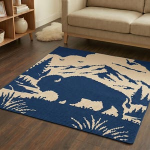 Puede incluir: Una alfombra rectangular con un dise&ntilde;o azul y beige que presenta una silueta de bisonte, monta&ntilde;as y &aacute;rboles. La alfombra est&aacute; colocada sobre un suelo de madera oscura, con un sof&aacute; beige y una estanter&iacute;a de madera en el fondo.