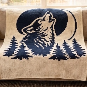 Op de afbeelding: Crèmekleurige gehaakte deken met een marineblauw ontwerp van een huilende wolf voor een volle maan en dennenbomen. De deken is over een beige bank gedrapeerd en toont het gedetailleerde vakmanschap en kleurcontrast.