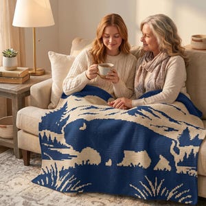 Puede incluir: Una manta de punto azul marino y crema con una silueta de bisonte en un dise&ntilde;o de paisaje. La manta est&aacute; sobre un sof&aacute; beige, con dos personas sentadas en &eacute;l. La escena est&aacute; ambientada en un sal&oacute;n acogedor.