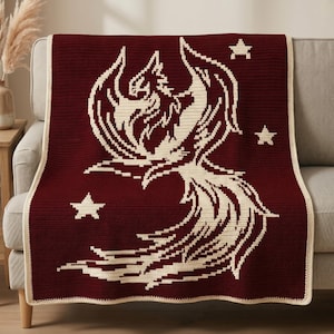May include: A rectangular crochet blanket in deep burgundy with a cream-colored phoenix design centered on the fabric. The phoenix is depicted with outstretched wings and a long, flowing tail, surrounded by three small cream-colored stars.
