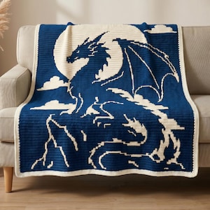 May include: A rectangular crochet blanket featuring a dark blue and cream color scheme. The design displays a large dragon silhouette perched on a rocky cliff against a full moon background, with small clouds scattered across the blue night sky.