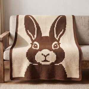Puede incluir: Una manta de ganchillo rectangular que presenta un gráfico grande y centrado de una cabeza de conejo. El diseño utiliza un esquema de dos colores, marrón oscuro y crema, con las orejas, los ojos y la nariz del conejo claramente definidos.