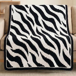 Peut inclure: Une couverture à rayures zébrées noires et blanches posée sur un canapé beige. La couverture présente un motif audacieux de rayures noires sur un fond blanc. Les bords sont finis par une bordure noire unie. Idéal pour la décoration intérieure.