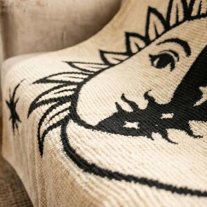 May include: Cream-colored throw blanket with a black sun and moon design. The celestial pattern is detailed with stars and a smiling face. The blanket is draped over a beige chair, showcasing its textured weave.
