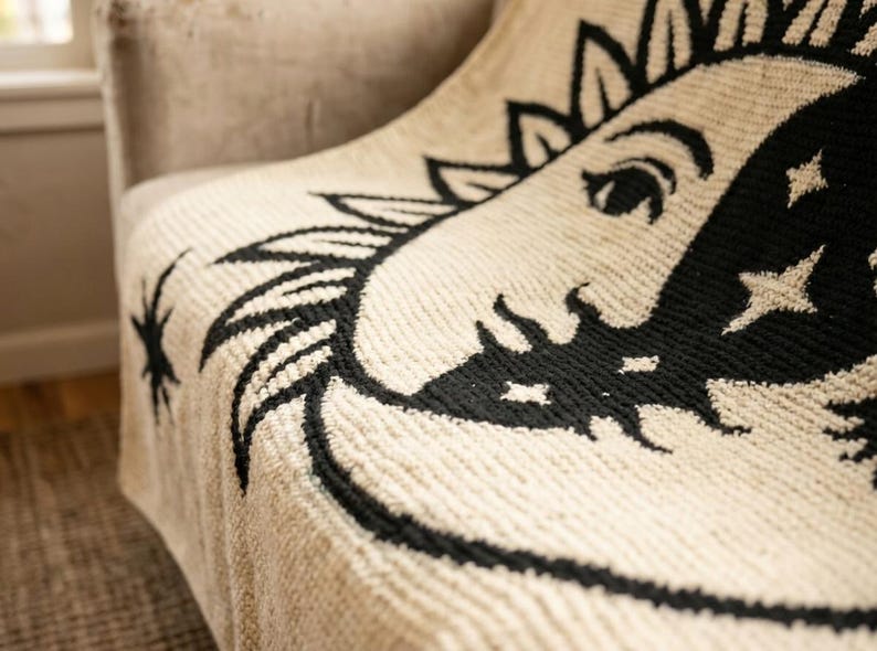 May include: Cream-colored throw blanket with a black sun and moon design. The celestial pattern is detailed with stars and a smiling face. The blanket is draped over a beige chair, showcasing its textured weave.