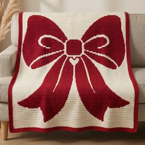 Puede incluir: Una manta de ganchillo rectangular que presenta un gran diseño de lazo rojo oscuro centrado sobre un fondo color crema. La manta tiene un borde sólido de color rojo oscuro que enmarca toda la pieza, con un pequeño corazón en el centro.