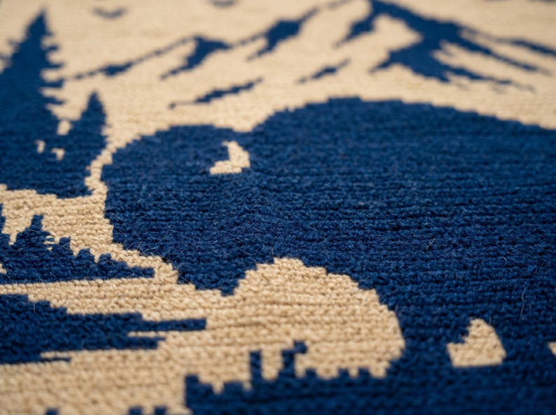 Puede incluir: Primer plano de una alfombra texturizada con un dise&ntilde;o de monta&ntilde;a y bisonte. Las siluetas de bisonte y monta&ntilde;a son de color azul marino sobre un fondo beige. La alfombra tiene una textura suave y tejida.