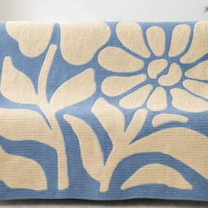 Puede incluir: Una manta de punto azul y crema con un gran diseño floral. La manta está sobre un sofá gris. El patrón floral presenta una flor grande y hojas en crema sobre un fondo azul.