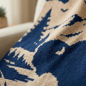 Puede incluir: Primer plano de una manta de ganchillo beige y azul marino. La manta presenta un dise&ntilde;o de monta&ntilde;a y &aacute;rboles. La manta est&aacute; colocada sobre un sill&oacute;n de color claro.
