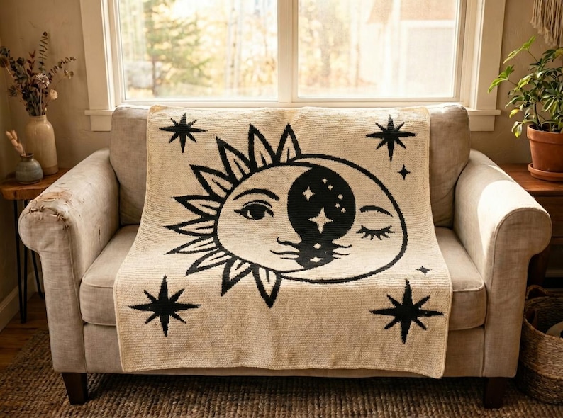 May include: A beige throw blanket draped over a sofa, featuring a black sun and moon design with stars. The blanket is made of a textured material and adds a decorative element to the living room. The sofa is a neutral color.