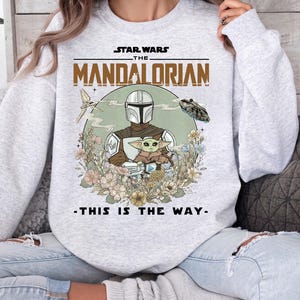 May include: A light gray crewneck sweatshirt featuring a graphic print of The Mandalorian and Grogu surrounded by flowers. The text reads STAR WARS THE MANDALORIAN and THIS IS THE WAY. A small spaceship is visible in the upper right corner.
