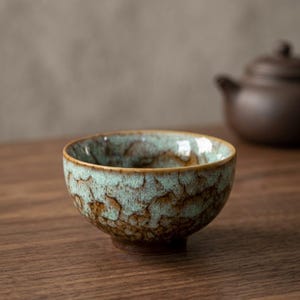May include: A small ceramic tea bowl featuring a mottled glaze with a blend of light mint green and deep brown tones. The exterior surface displays a crackled texture with dark brown accents near the base, transitioning into a lighter green.