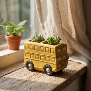 May include: A yellow ceramic planter shaped like a double-decker bus with two open compartments on the roof. The planter holds a green succulent plant in each section, featuring molded windows, black wheels, and a matte finish on the bus body.