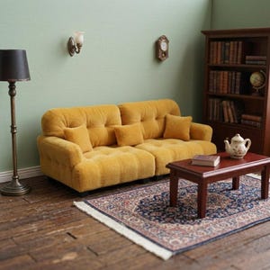 May include: A miniature mustard yellow tufted sofa with three matching square throw pillows. The sofa is positioned in a room setting featuring a dark wood coffee table, a floor lamp, a wooden bookshelf, and a patterned area rug on a wood floor.