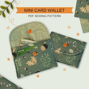 May include: Two mini card wallets made from green fabric with an acorn and leaf print. One wallet is open, showing a light green polka dot interior lining and a credit card inside. Both wallets feature a small orange snap button closure.