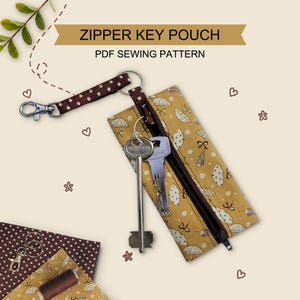Peut inclure: Une pochette à clés rectangulaire en tissu jaune moutarde avec un motif de parapluies blancs et une fermeture éclair marron foncé. Une sangle à pois marron et blanc avec un mousqueton en métal est fixée à un anneau portant deux clés.
