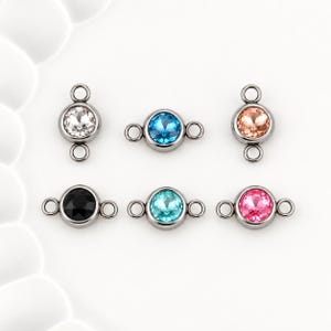 May include: Six circular jewelry connectors featuring faceted crystals in clear, blue, peach, black, turquoise, and pink. Each connector is set in a dark metal bezel with two small loops on opposite sides for attaching to chains or other jewelry components.