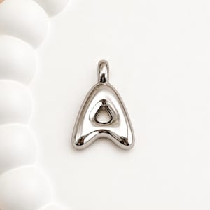 May include: A silver-toned metal pendant shaped like the capital letter A. The design features a smooth, rounded, puffed surface with a reflective finish and a small triangular cutout in the center. A small loop is attached to the top for hanging.