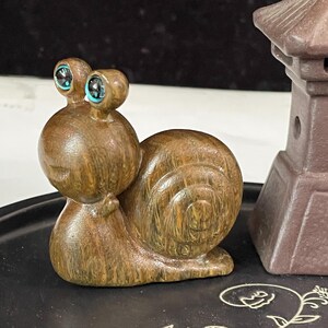 Può includere: Una statuetta di lumaca in legno marrone con finitura lucida e due occhi prominenti caratterizzati da centri circolari blu brillante. La lumaca presenta un guscio arrotondato con venature del legno visibili ed è posizionata accanto a una lanterna in pietra.