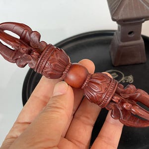 May include: A dark reddish-brown wooden vajra ritual object featuring intricate symmetrical carvings of lotus petals and curved prongs on both ends, centred by a smooth spherical bead, showcasing detailed craftsmanship and a polished finish.
