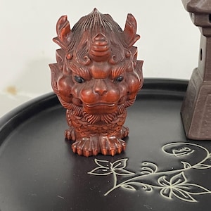 Puede incluir: Una detallada escultura de madera marrón rojizo de una criatura mítica con rostro de león, cuernos y tallas intrincadas. La figura está sobre una superficie negra con un diseño floral. Una pequeña estructura marrón con forma de pagoda es visible en el fondo.