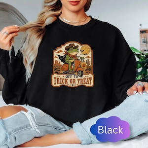 May include: A black crewneck sweatshirt featuring a graphic of a green frog wearing a cowboy hat and cape, riding a scooter through a desert landscape. The design includes a full moon, cacti, and the text "OFF TO TRICK OR TREAT" in a bold font.