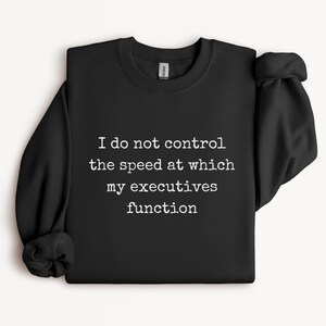 May include: A black long-sleeve crewneck sweatshirt featuring white typewriter-style text on the front that reads, I do not control the speed at which my executives function. The garment has a ribbed collar, cuffs, and hem with a standard fit.