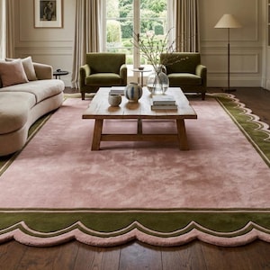 Op de afbeelding: Een groot, rechthoekig vloerkleed met een geschulpte rand. Het vloerkleed heeft een zachtroze midden, omzoomd door een donkergroen geschulpt ontwerp. Het vloerkleed ligt in een woonkamer met een gebogen beige bank en groene fauteuils.
