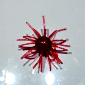 May include: A translucent red plastic fishing lure featuring a central rounded body with numerous thin, radiating tentacles extending outward in a starburst pattern, designed to mimic the appearance of a small aquatic organism or crustacean.