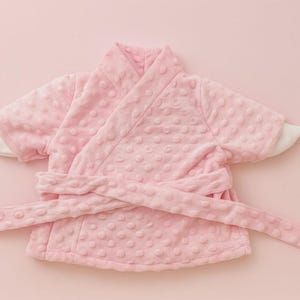 May include: A light pink baby robe with a textured, dotted pattern. The robe has a wrap-around design with a matching belt and white cuffs. The soft, plush material suggests comfort and warmth. The robe is designed for infants or toddlers.