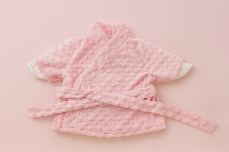 May include: A light pink baby robe with a textured, dotted pattern. The robe has a wrap-around design with a matching belt and white cuffs. The soft, plush material suggests comfort and warmth. The robe is designed for infants or toddlers.