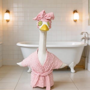 May include: A whimsical sculpture of a goose in a spa setting. The goose wears a pink terry cloth robe and a matching head wrap with cucumber slices over its eyes. The background features a white bathtub and tiled walls, creating a relaxing spa theme.