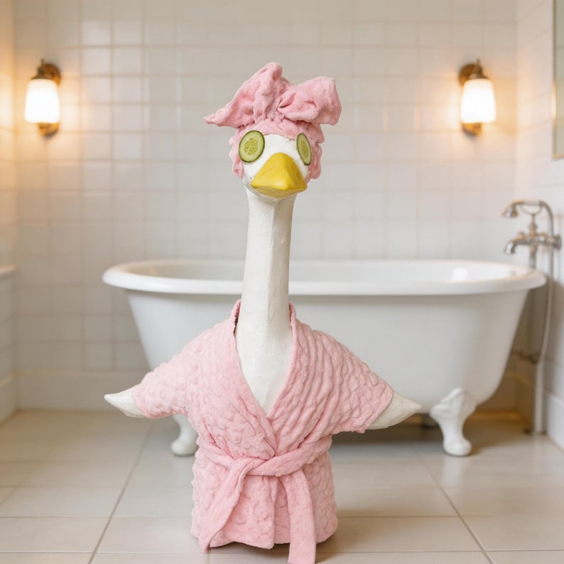 May include: A whimsical sculpture of a goose in a spa setting. The goose wears a pink terry cloth robe and a matching head wrap with cucumber slices over its eyes. The background features a white bathtub and tiled walls, creating a relaxing spa theme.