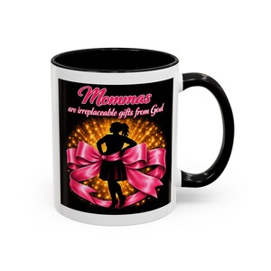 May include: White ceramic mug with a black interior and handle. The mug features a design with the text "Mommas are irreplaceable gifts from God" above a silhouette of a woman in a pink bow.