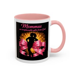 May include: White ceramic mug with a pink interior and handle. The mug features the text "Mommas are irreplaceable gifts from God" above a silhouette of a woman and a large pink bow on a black background.