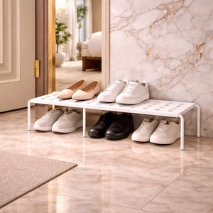 May include: A white metal shoe rack holding various pairs of shoes. The shoes include beige flats, white sneakers, gray sneakers, and black dress shoes. The rack is placed in a well-lit hallway with a marble-patterned wall.