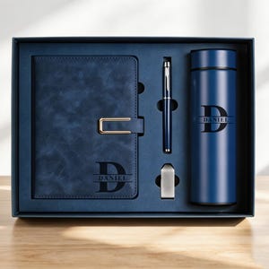 May include: A navy blue gift set in a box, including a journal, pen, bottle, and a small silver item. The journal and bottle are personalized with the name "DANIEL". The journal has a gold clasp. The pen is silver and navy blue.