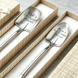 May include: Two silver-toned metal ice cream shovels with long handles, each resting in a beige plaid gift box. One shovel is engraved with the text "Dad's Ice Cream Shovel" and the other with "Mom's Ice Cream Shovel" accompanied by a small ice cream cone icon.