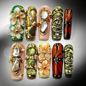 May include: A set of ten press-on nails featuring intricate 3D designs. The nails display various patterns including sculpted flowers, gold metallic swirls, iridescent shell-like inlays, butterfly motifs, and glossy orange and black wing patterns.