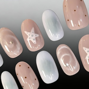 Puede incluir: Un conjunto de diez uñas postizas ovaladas que presentan una mezcla de blanco sólido, rosa nude con puntos dorados metálicos, rosa nude con brillo de ojo de gato y una pequeña estrella plateada, y rosa nude con un diseño de estrella de mar.