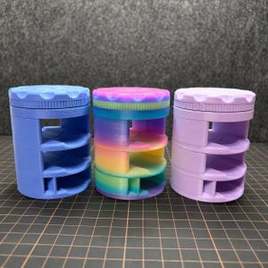 Puede incluir: Tres contenedores de almacenamiento cilíndricos en azul, degradado arcoíris y lavanda. Cada uno tiene una tapa de rosca y tres estantes. Los contenedores son de plástico y están diseñados para objetos pequeños.