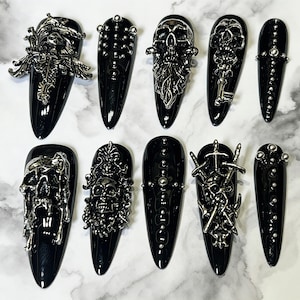 Peut inclure: Un ensemble de dix faux ongles en forme de stylet noir brillant, ornés de breloques métalliques argentées en trois dimensions, notamment des crânes, des épées, des filigranes ornés et de petits clous métalliques disposés en motifs verticaux.