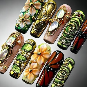 Puede incluir: Un conjunto de diez uñas postizas largas con diseños 3D intrincados. Las uñas muestran una mezcla de purpurina verde, remolinos metálicos dorados, acentos iridiscentes tipo concha, pequeñas perlas blancas y pétalos de flores de color naranja.