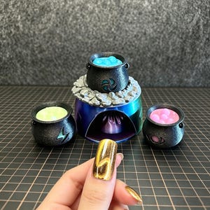 Puede incluir: Colección de calderos negros en miniatura con contenidos coloridos, incluyendo azul, verde y rosa. Los calderos están dispuestos alrededor de un soporte decorativo azul y plateado con un diseño de llama. La imagen tiene un tema de brujería o fantasía.