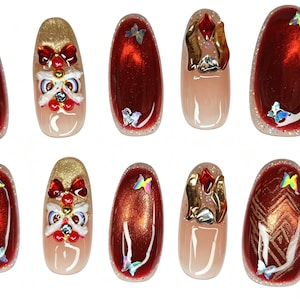 May include: A set of ten press-on nails featuring a deep red cat-eye polish base with gold glitter accents. Decorations include iridescent butterfly charms, red bows, white lion-like face ornaments, and metallic gold geometric patterns.