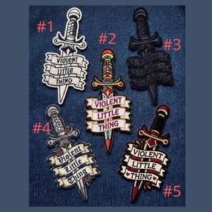 May include: Embroidered patches featuring dagger designs with the text "VIOLENT LITTLE THING" in various styles. The patches are in black, white, red, and gold colors, with detailed stitching. The patches are displayed on a blue denim background.