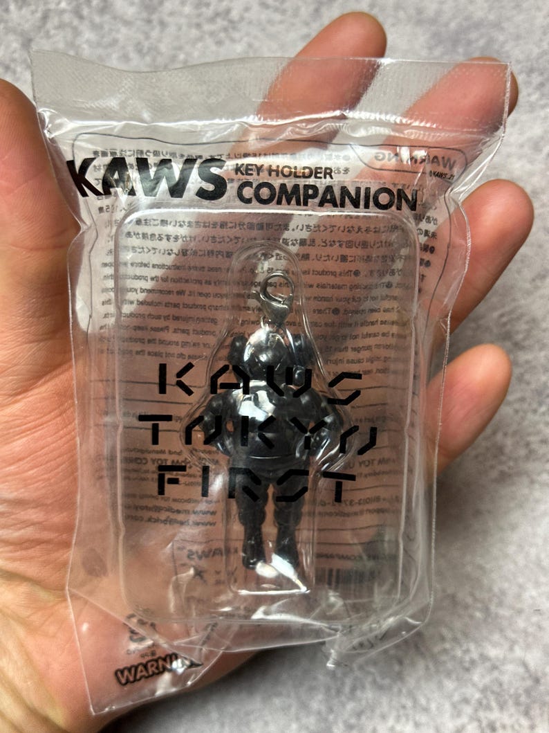 May include: A black KAWS Companion key holder figure in a clear plastic package. The package has the text "KAWS KEY HOLDER COMPANION" in black. The figure is in a standing pose with arms at its sides. The figure has a silver key ring.